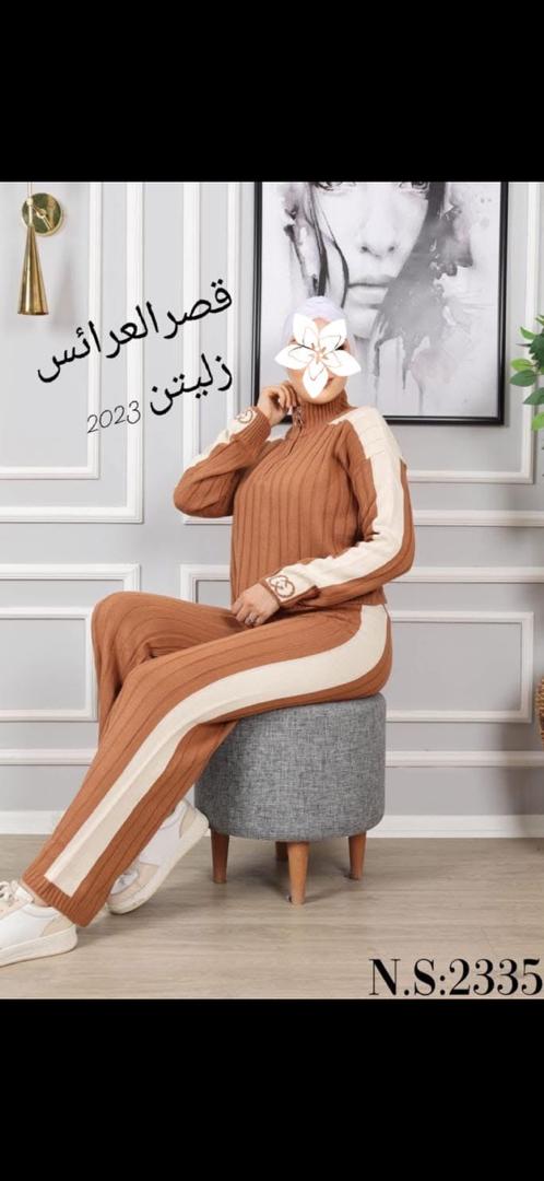 طقم نسائي محبوك بني غامق مكون من بلوزة بياقة عالية وسروال واسع مع شريط كريمي على الجانبين، المرأة ترتدي حجاب أبيض ووجهها مغطى بملصق زهرة، جالسة على مقعد رمادي في استوديو أمام حائط مزخرف وإطار لوحة، نص داخل الصورة قصر العرائس زليتن 2023 ورقم المنتج N.S:2335، انطباع أنيق ومريح ومحافظ