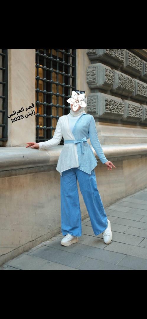 عارضة ترتدي حجاب أبيض وسترة بلونين أزرق فاتح وبيج وبنطلون جينز واسع تقف على رصيف أمام واجهة حجرية مزخرفة الوجه مغطى بملصق زهرة نص داخل الصورة قصر العرائس زليتن 2025 الانطباع أنيق وواثق