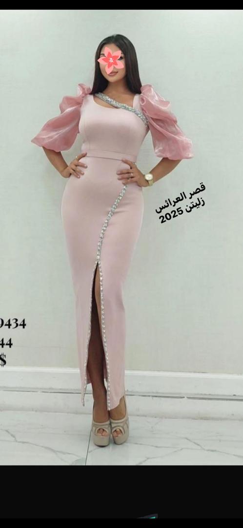 امرأة ترتدي طقماً أنيقاً مكوناً من بلوزة وسروال، تقف في متجر ملابس مع خلفية تعرض مجموعة متنوعة من الأزياء. النص الموجود في الصورة يقرأ قصر العرائس زليتن 2026، مما يضفي طابعاً عصريًا وثقة على المشهد.